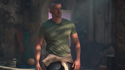 A man in a t-shirt stands in the shadows looking up at something off screen