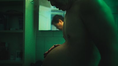 A man holding a rounded belly - the image is taken from side profile and is quite dark with a green hue