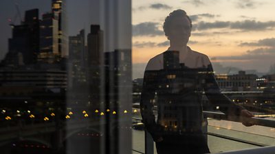 A man is reflected in glass, standing on a balcony along the River Thames in London, with buildings and lights on a bridge also reflected in the pane