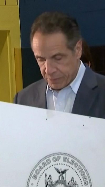 A man wearing a grey suit looks down as he participates in voting, his lower half hidden by a polling separator.