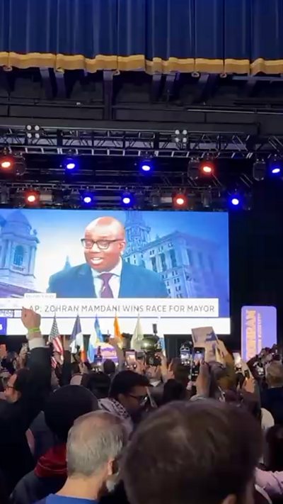 A crowded room before a screen onstage showing Mamdani's win.