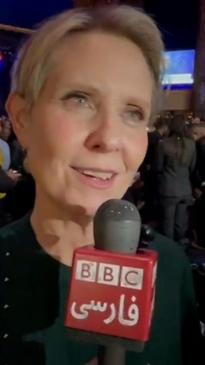 Cynthia Nixon wears a green shirt as she speaks into a red BBC microphone with a large crowd behind her.