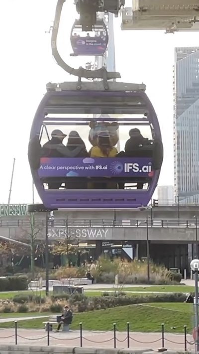Pudsey sits in a purple cable car gliding above a park and walkway.
