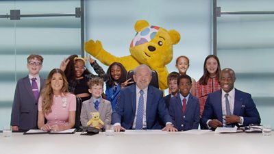 Lord Sugar, Karen Brady and Tim Campbell sit at the Boardroom table smiling, surrounded by eight children and Pudsey. There are papers and glasses of water on the desk in front of them.