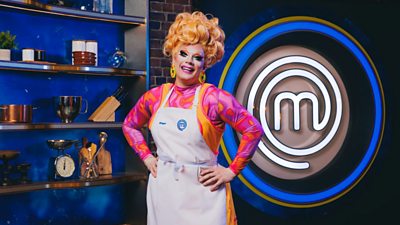 Ginger Johnson wearing a white apron in the exposed brick MasterChef kitchen. Cooking equipment lines the shelves behind her and a large MasterChef logo in lights is to her left.
