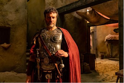 A man, in Ancient Rome dress, stands proudly with his armour. 