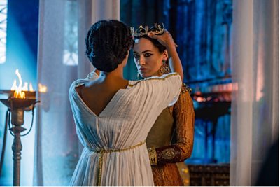 A woman in Ancient Egypt is crowned by another woman in regal dress, with a fire burning in the background.