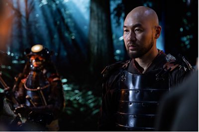 A Japanese man stands in the forest, in full Samurai armour. 
