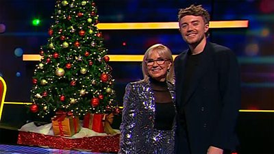 A woman in a glittery suit and a man in a dark suit stand together on set smiling beside a Christmas tree