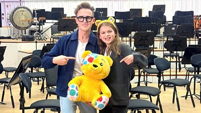 A man in a blue shirt, white t-shirt, jeans and glasses and a boy in a grey cardigan wearing Pudsey Bear ears stand together smiling holding a large yellow soft toy Pudsey Bear. They pose in front of a selection of empty orchestra chairs.