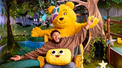 A man in a brown hoodie sits in a chair with his arms thrown wide, smiling. Pudsey Bear stands behind him with his arms spread wide also. The pair pose against a background designed to look like a magical forest.