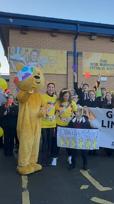 Pudsey, Amy Garcia and Lindsey Burrow have arms around each other while children wave flags behind them.