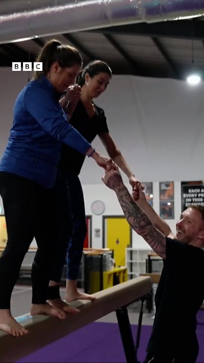 Two women stood on a balance beam holding a man's hands for balance