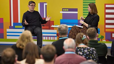 Greater Manchester mayor Andy Burnham sits on a stage alongside BBC reporter Alex Forsyth. Mr Burnham has an open pose as he answers a question