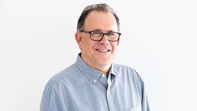 A man with a blue checked shirt, grey hair and glasses smiles at the camera