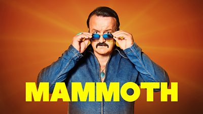 A man in a denim jumpsuit looks to camera, lowering a pair of sunglasses