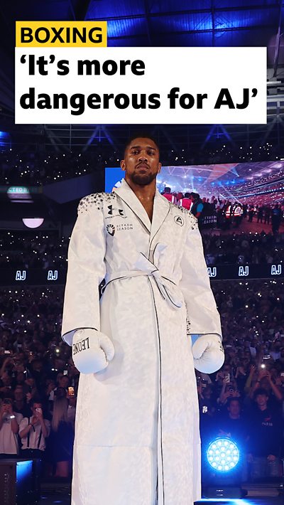 Boxer Anthony Joshua wearing a white robe and boxing gloves. He is standing in the ring in Wembley Stadium.