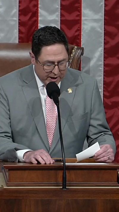 Man wearing a gray suit speaks behind a podium with an American flag behind him.