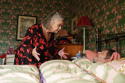A woman stands over a man who's lying in bed. She looks angry and stands with her hands outstretched in a frustrated gesture while he looks up at her.