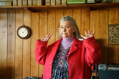 A woman in a red coat, patterned dress and purple jumper stands with her hands up in a room with wood panelling on the walls