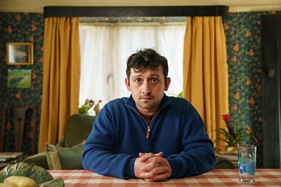 A man in a blue zip neck jumper sits at a table looking worried
