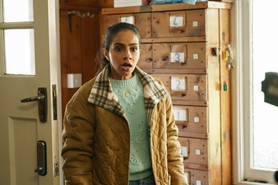 A woman in a beige coat and green jumper walks in to a room, looking shocked by something she sees off camera