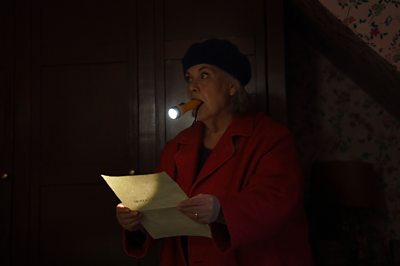 A woman in a red coat and dark hat stands in the dark with a torch in her mouth, holding a piece of paper and looking wary