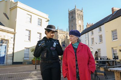 A woman in police uniform and a woman in a red coat and purple hat walk side by side down a street with a church and houses visible in the background