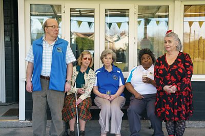 A woman in a black and red patterned dress perches on the end of a bench she shares with three women and a man in a sleeveless fleece stands at the other end.
