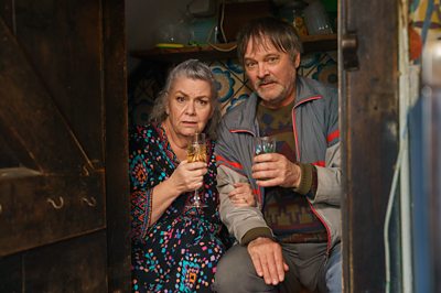 Two people huddle together in a wooden cupboard, holding drinking glasses and looking a bit sheepish