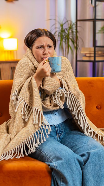 A woman wrapped in a blanket, holding a mug while sat on a sofa at home