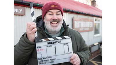 A man in a warm coat and red hat stands outside a large metal shed with a red roof, holding a clapperboard for the start of filming on Shedites.