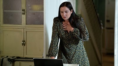 Lauren stood at a kitchen table looking horrified at a laptop in front of her. Her hand is pressed to her chest in shock.