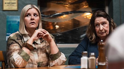 Two women sat at a tabble in a cafe at night looking at something out of shot off to their right
