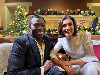 A man and a woman sit together on a sofa smiling with Christmas decorations in the background 