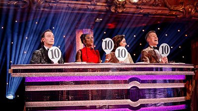 Craig Revel Horwood, Motsi Mabuse, Shirley Ballas and Anton Du Beke sit behind a glittering desk in the Blackpool Tower Ballroom in their finery, each holding a glittery 10 paddle