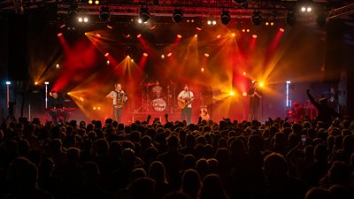 Photo of Trail West performing onstage to a large crowd of concertgoers. 