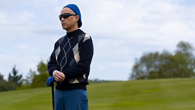 Photo of Ken Leung in character as Eric Tao. Wearing a patterned jumper, a blue hat, golfing glove and trousers. He leans on a golf club and watches something off in the distance. 