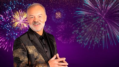 A man in a black suit jacket with a gold design smiles to camera, set against a purple background featuring fireworks