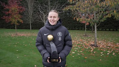 A young woman stood outside on some grass next to some small trees in Autumn, holding the BBC Women’s Footballer of the Year 2025 trophy which is made up of glass and a golden football. She is smiling.