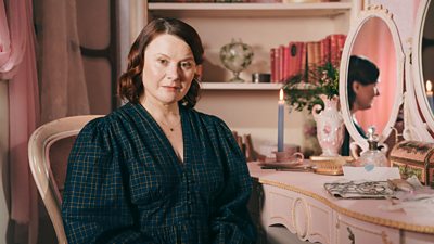 A woman in a patterned dark green/blue dress sits at a dressing table, her face reflected in the mirror on the vanity and a single candle lit on the dressing table