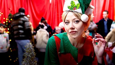 A woman dressed as one of Father Christmas' elves stares to camera, slightly smirking, as parents and children bustle around behind her