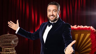 A man in a dark velvet suit and bow tie smiles as he extends his arms outwards, ready for start a show