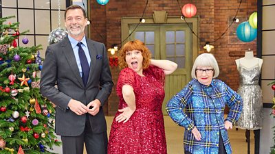 A man in a suit, a woman in a sparkling red dress and a woman in a blue patterned coat style dress each strike a pose in a sewing room