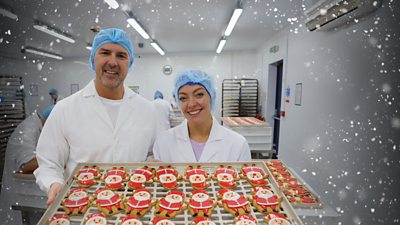 A man and woman in white coats and blue caps smile as they pose with a large thread of gingerbread Santas, brightly decorated with red and white icing