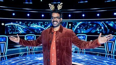 A man in a dark brown shirt jacket, orange shirt and reindeer antlers smiles as he holds his arms out on the set of a quiz show