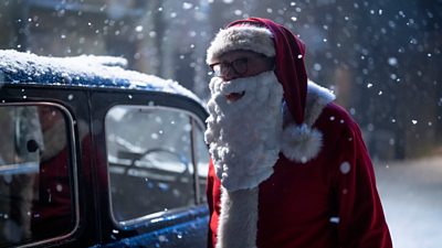 A man dressed as Father Christmas in the snow stood next to an old fashioned blue car
