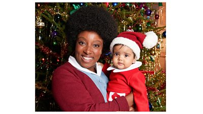 A woman smiling, wearing a blue and white nurse's dress and red cardigan, holding a baby dressed in a Santa hat, in front of a decorated Christmas tree
