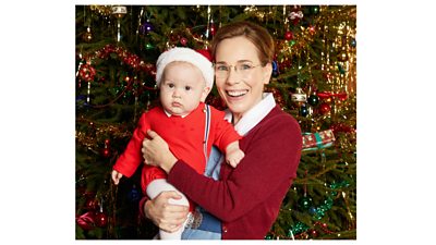 A woman smiling, wearing a blue and white nurse's dress and red cardigan, holding a baby dressed in a Santa hat, in front of a decorated Christmas tree