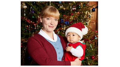 A woman smiling, wearing a blue and white nurse's dress and red cardigan, holding a baby dressed in a Santa hat, in front of a decorated Christmas tree